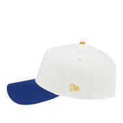 Side view of a New Era 9FORTY A-Frame cap with a white crown and royal blue brim, featuring a gold embroidered New Era flag logo on the side panel.