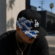 Male wearing a black Los Angeles Dodgers snapback hat with blue Japanese wave embroidery and white LA logo in an urban streetwear setting.