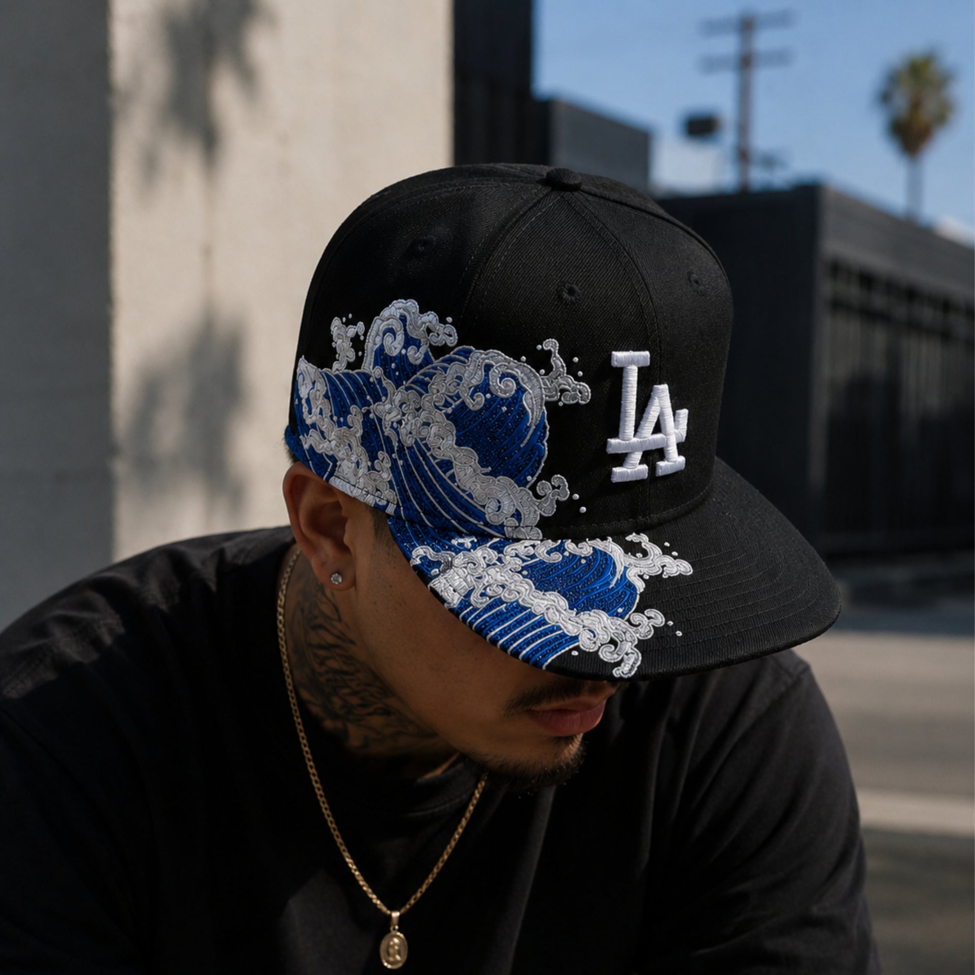 Male wearing a black Los Angeles Dodgers snapback hat with blue Japanese wave embroidery and white LA logo in an urban streetwear setting.