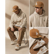 Person wearing a beige hoodie, brown pants, and white sneakers with a LA cap, sitting on a chair.