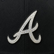 A close-up of a black baseball cap featuring a large, silver rhinestone-embellished script letter 'A' on the front panel.