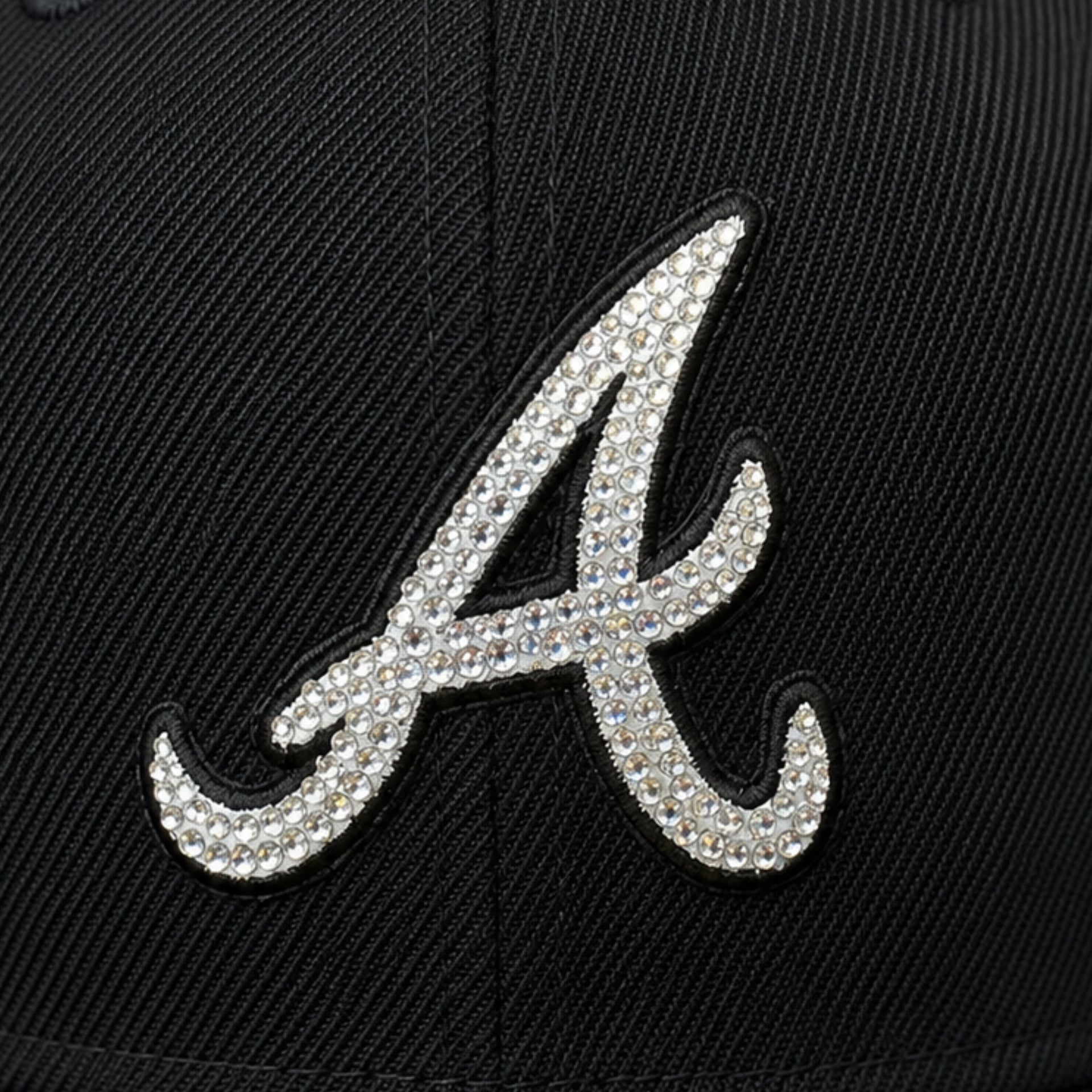 A close-up of a black baseball cap featuring a large, silver rhinestone-embellished script letter 'A' on the front panel.