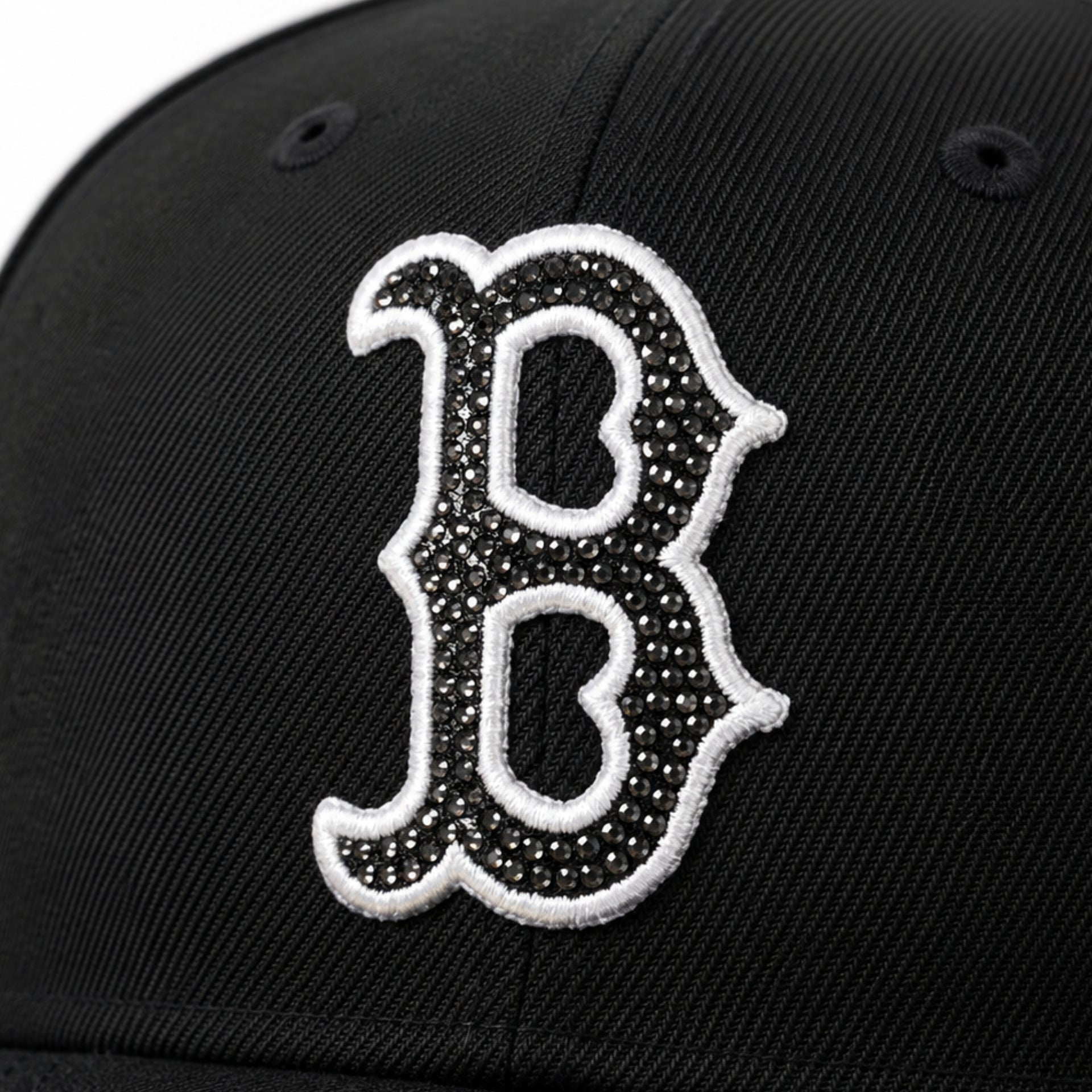 Close-up of a black baseball cap featuring a large embroidered Boston Red Sox letter 'B' logo embellished with rhinestones and outlined in white.