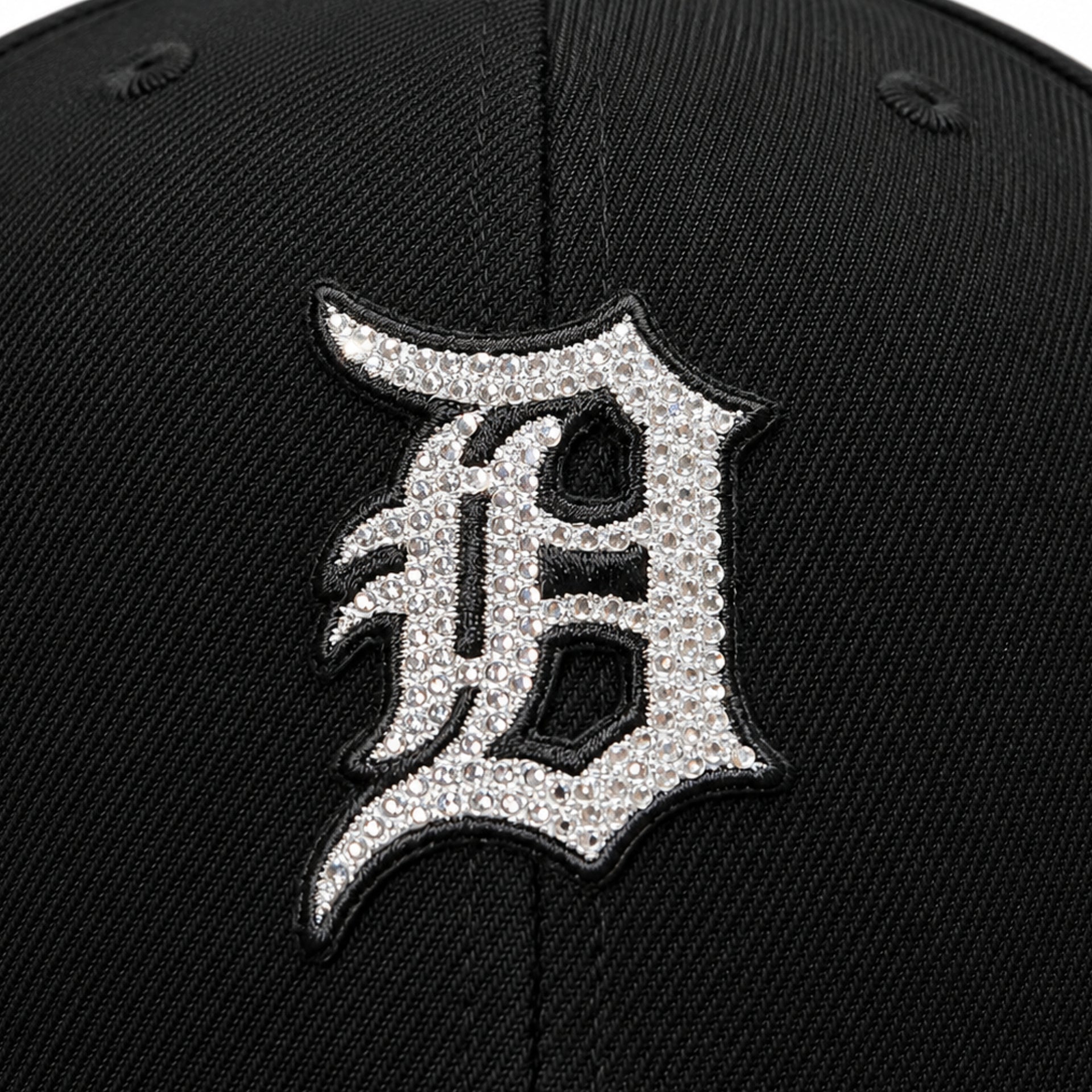 Close-up of a black baseball cap featuring a large embroidered letter 'D' patch covered in silver rhinestones, outlined in black.