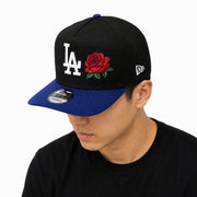 a male wearing New Era Los Angeles Dodgers Rose 9FIFTY A-Frame Snapback Hat in black and royal blue with embroidered LA logo and red rose detail.