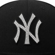 Close-up of a black baseball cap featuring a large New York Yankees 'NY' logo patch embellished with silver rhinestones.