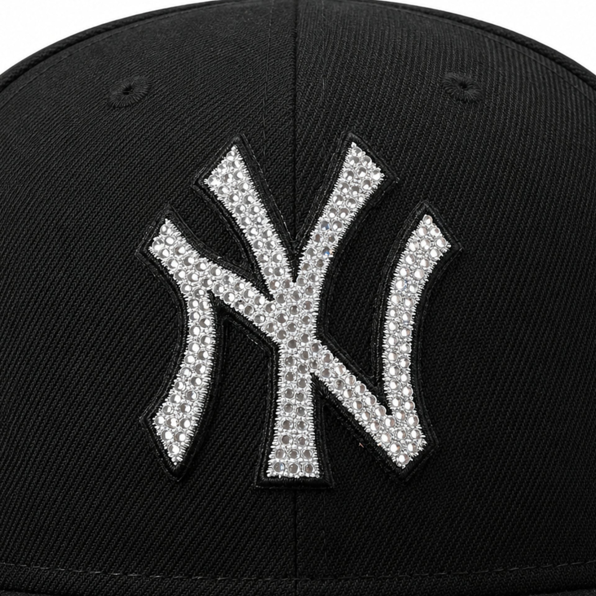 Close-up of a black baseball cap featuring a large New York Yankees 'NY' logo patch embellished with silver rhinestones.